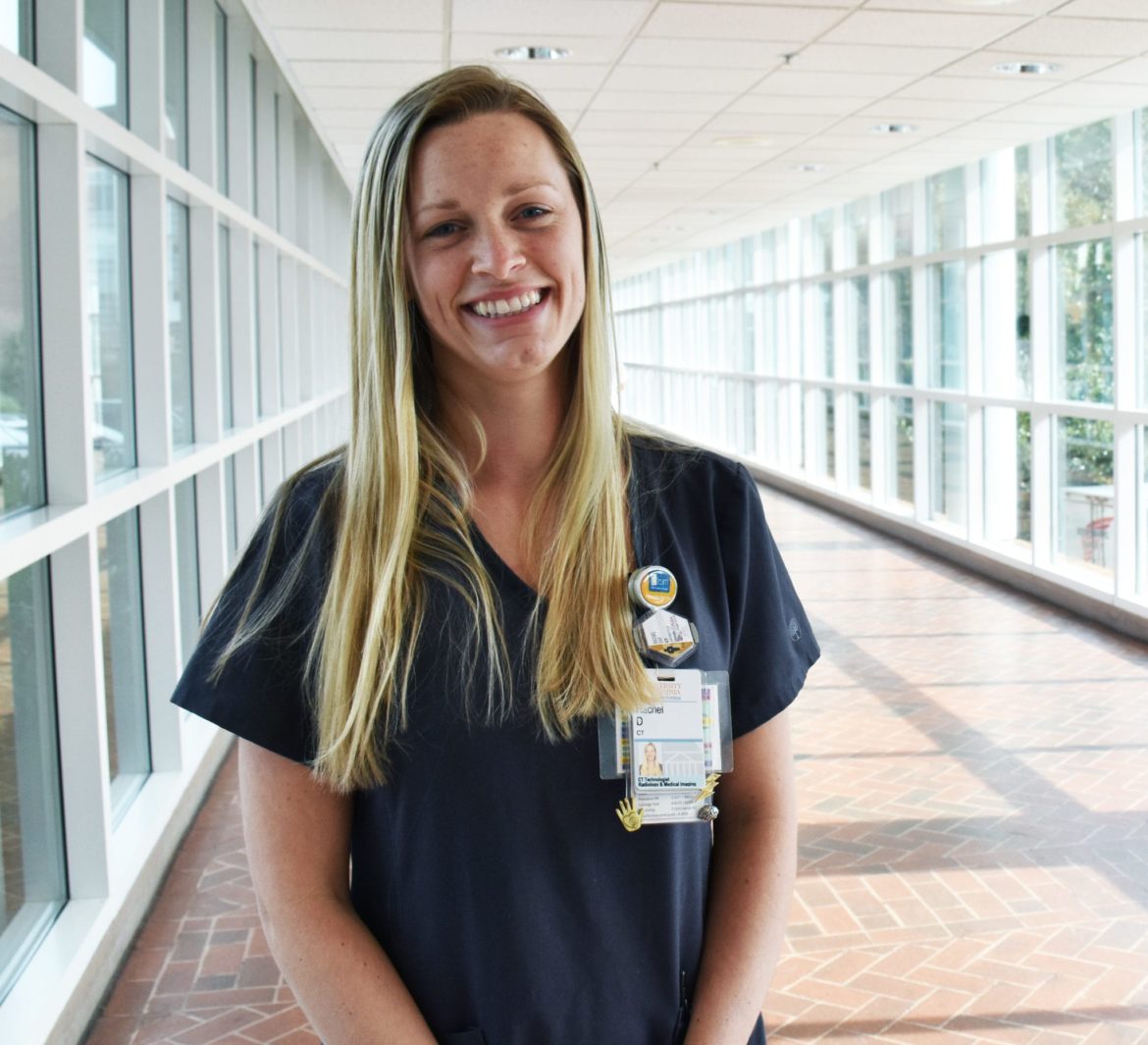 Woman CT technologist, Rachel Day, stands in a well lit hallway and smiles at the camera