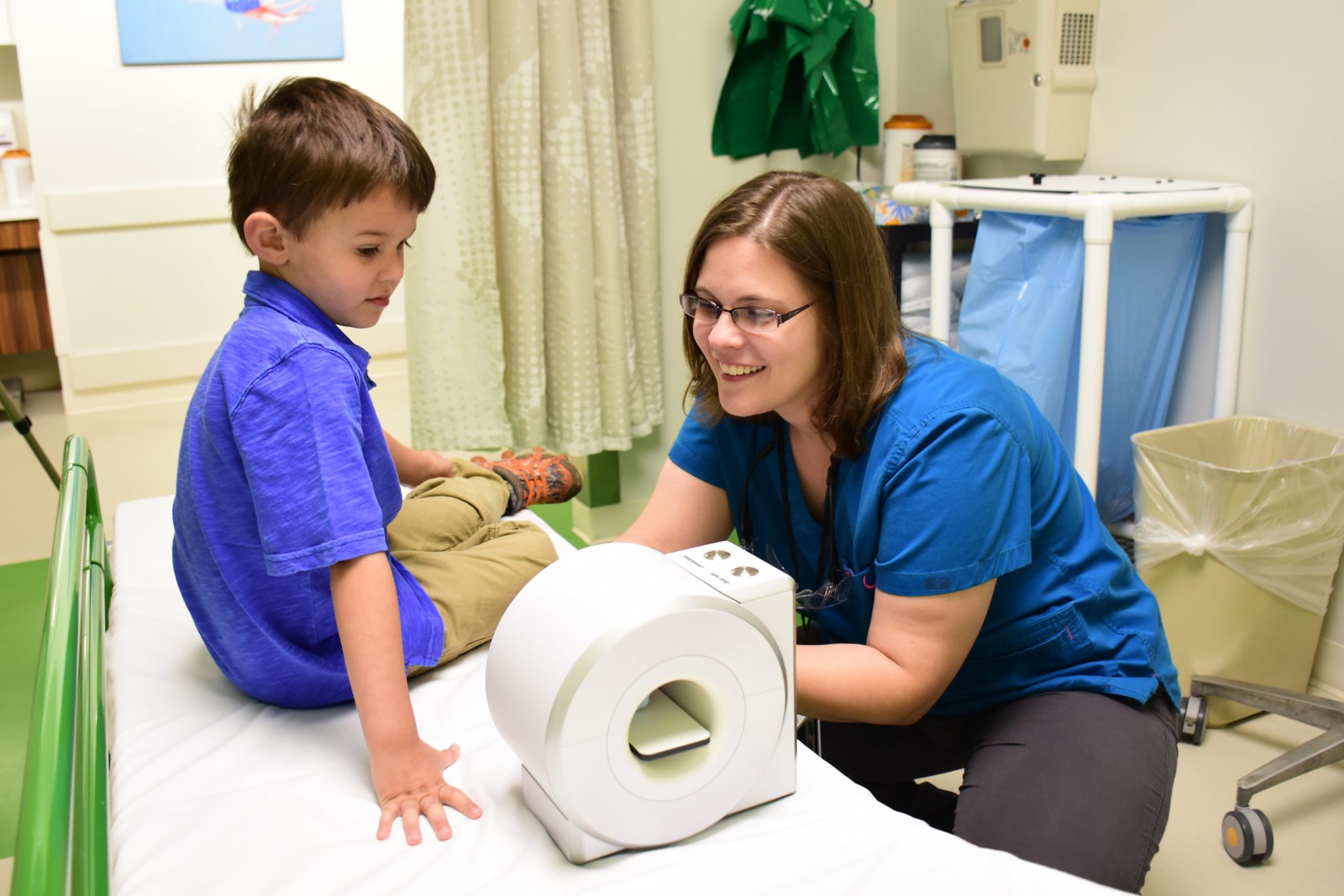 A boy sits in a waiting room as a technician explains how an MRI works using a toy man and a toy MRI