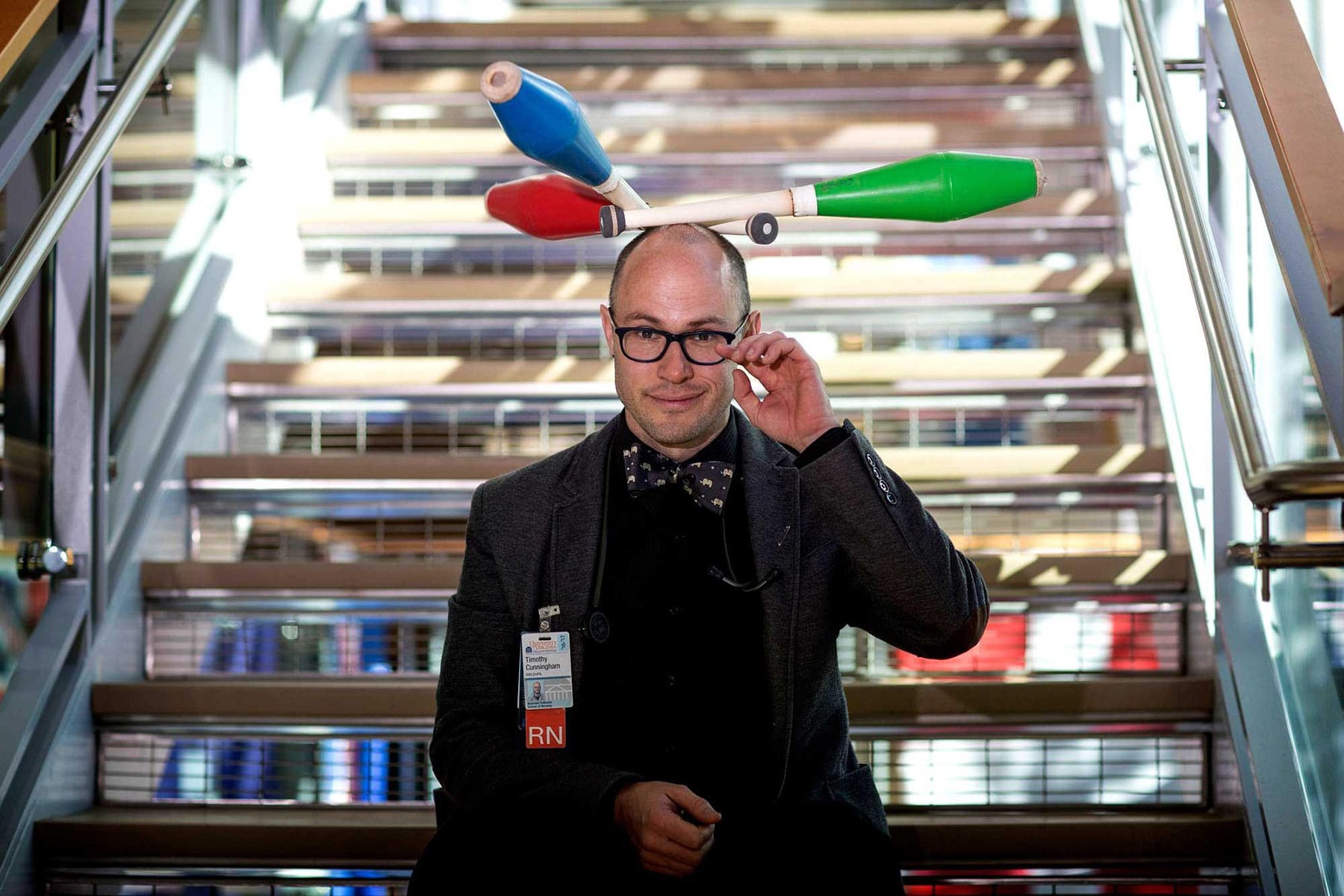 Tim Cunningham, a former clown, now a nurse, sits on a glass staircase with three juggling pins balanced on his head.